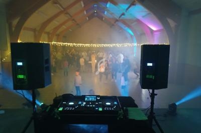 Vue depuis une table de mixage avec des enceintes, éclairage coloré et foule dansante dans une salle festive.