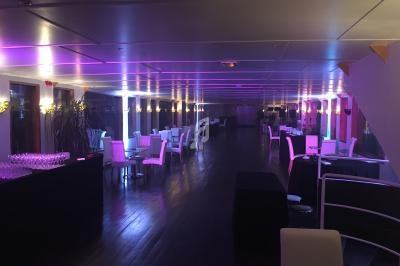 Salle de réception moderne avec éclairage violet, tables dressées et chaises blanches sur un sol en bois sombre.