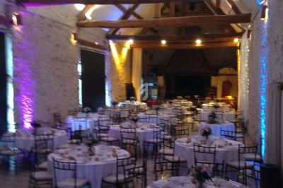 Salle de réception avec poutres apparentes, tables rondes dressées pour un événement et éclairage coloré.