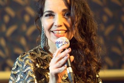 Femme souriante en robe dorée tenant un micro, devant un fond sombre avec des motifs.