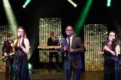 Des chanteurs et musiciens en tenue de soirée se produisent sur une scène éclairée par des projecteurs colorés.