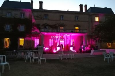 Façade d'un bâtiment éclairée par des lumières roses au crépuscule, avec des chaises et tables disposées sur la pelouse.