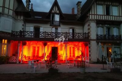 Façade d'une grande maison à colombages éclairée par des lumières rouges au crépuscule, avec mobilier de jardin.