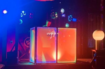 Un DJ mixe derrière une cabine lumineuse entourée de jeux de lumière colorés dans une ambiance festive.