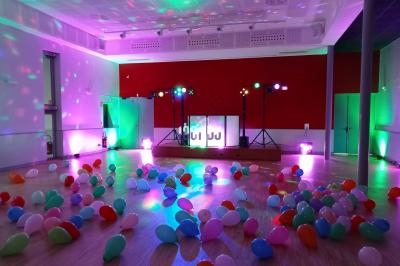 Salle de fête décorée avec des ballons colorés au sol et des lumières d'ambiance projetées sur les murs.