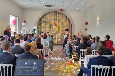 Cérémonie de mariage en intérieur avec invités assis, décorations festives et une personne lisant un discours.