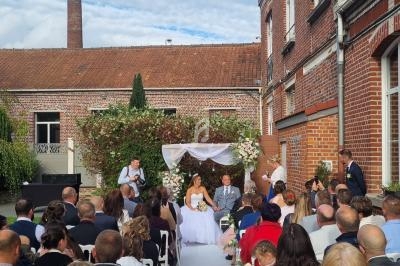 Cérémonie de mariage en extérieur avec des invités assis, un couple sous une arche fleurie et un ciel partiellement nuageux.