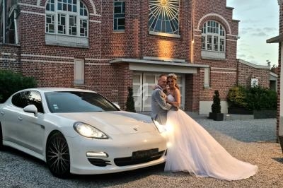 Un couple en tenue de mariage pose devant une voiture blanche et un bâtiment en briques au crépuscule.
