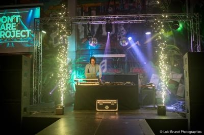 Un DJ mixe sur une scène éclairée par des projecteurs colorés et des étincelles, entouré de plantes et d'équipements.