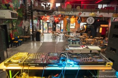 Poste de DJ équipé de platines et d'un ordinateur, installé dans une salle colorée avec tables et chaises en arrière-plan.