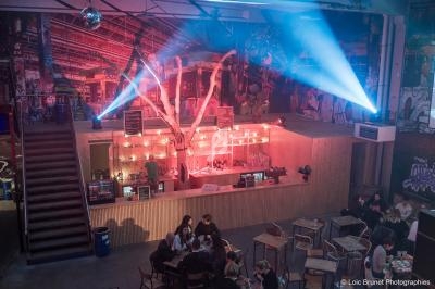 Intérieur d'un bar avec un comptoir éclairé, des tables occupées et des projecteurs illuminant le plafond.