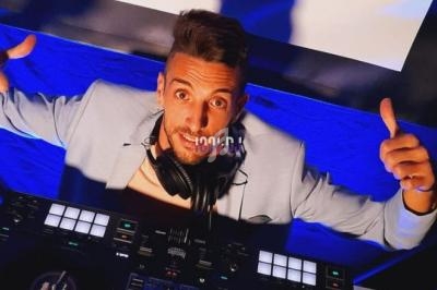 Un DJ souriant en costume clair mixe sur une table de mixage, éclairé par des lumières bleues.