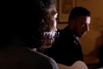 Deux personnes dans une pièce faiblement éclairée, l'une jouant de la guitare, l'autre de dos en premier plan.