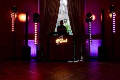 Un DJ mixe derrière une table éclairée par des lumières colorées dans une salle sombre avec de grandes fenêtres.