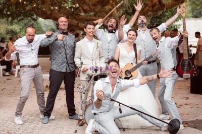 Un groupe de musiciens et une mariée posent joyeusement avec des instruments sous des arbres décorés de guirlandes…