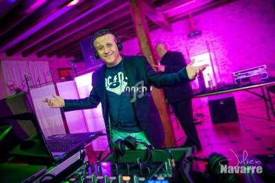 Un DJ souriant anime une soirée dans une salle éclairée par des lumières violettes, avec une table de mixage au premier plan.