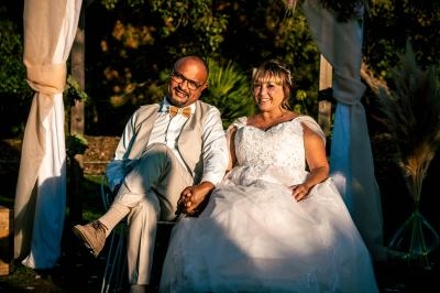 Un couple assis sous une arche décorée, souriant lors d'une cérémonie en extérieur par temps ensoleillé.
