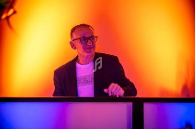 Un homme souriant en veste sombre se tient derrière un pupitre, éclairé par des lumières orange et bleues.