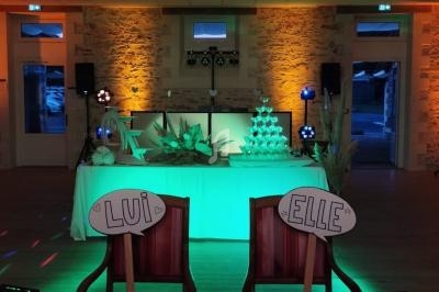 Salle décorée pour un événement, avec une table ornée de plantes, une pyramide de verres et deux chaises marquées ’Lui’ et…