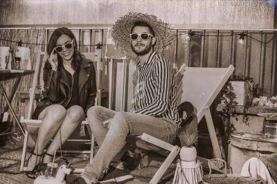 Un homme et une femme assis sur des chaises longues, portant des lunettes de soleil, dans un décor rétro extérieur.