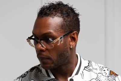 Homme portant des lunettes et une veste blanche à motifs, regardant vers sa gauche dans un environnement intérieur.