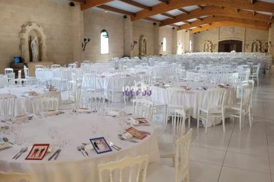 Salle de réception lumineuse avec tables rondes dressées, nappes blanches et chaises en bois blanc, sous un plafond en…