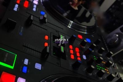 Table de mixage avec platines, boutons lumineux et écrans affichant des pistes audio en arrière-plan.