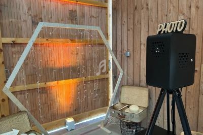 Cadre hexagonal décoré de guirlandes lumineuses, valises vintage et borne photo dans un espace en bois.