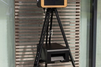 Borne photo sur trépied avec boîtier en bois, anneau lumineux et imprimante intégrée, posée devant un mur à lames.