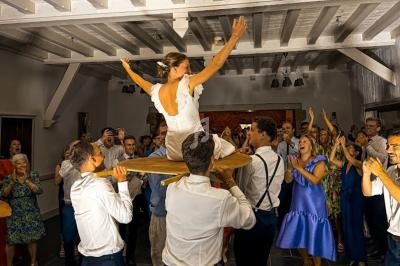 Une mariée est soulevée sur une chaise par des invités dans une salle festive, entourée de personnes applaudissant.