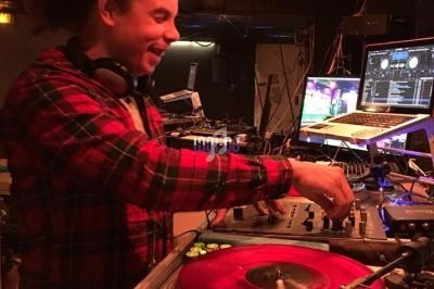 Un DJ souriant mixe de la musique sur des platines avec un vinyle rouge dans un environnement sombre.