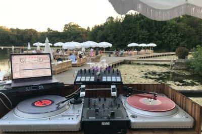 Poste de DJ en extérieur avec platines vinyles, ordinateur et table de mixage, vue sur un lac et une terrasse animée.