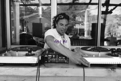 Un DJ mixe de la musique sur des platines vinyles, installé derrière une table en bois dans un environnement extérieur.