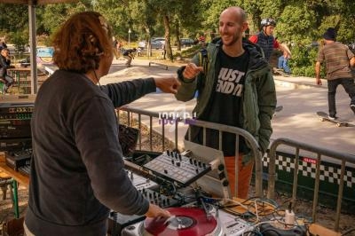 Un DJ échange un geste amical avec un homme près d'une table de mixage, dans un parc avec des skateurs en arrière-plan.