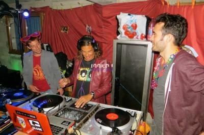 Trois personnes dans une pièce décorée, dont une manipulant des platines DJ, entourée de matériel audio.