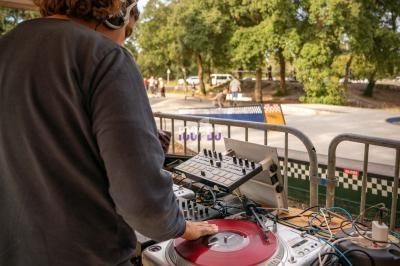 Un DJ mixe des vinyles sur une table de mixage en plein air, près d'un skatepark entouré d'arbres.