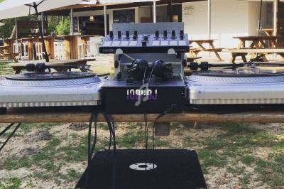 Deux platines vinyles et une table de mixage installées en extérieur, sous des parasols, près de tables en bois.