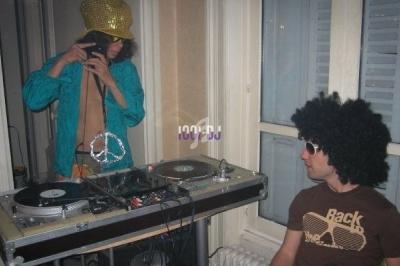 Deux personnes en tenue rétro, l'une mixant des disques vinyles sur une table de DJ, l'autre assise avec une perruque afro.