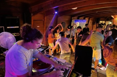 Un DJ mixe de la musique dans une salle en bois tandis que des personnes dansent sous des lumières colorées.