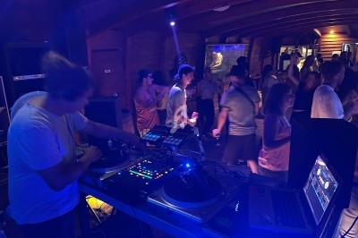 Un DJ mixe de la musique sur une table de mixage dans une salle sombre, entouré de personnes dansant.