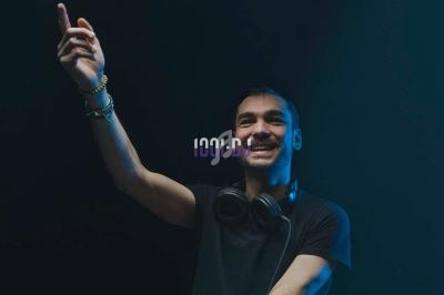 Un DJ souriant portant un casque audio autour du cou, levant une main dans une ambiance lumineuse sombre.