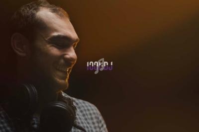 Un homme souriant portant un casque audio autour du cou, éclairé par une lumière chaude sur un fond sombre.