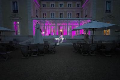 Façade d'un bâtiment éclairée en rose à la tombée de la nuit, avec terrasse aménagée de tables et parasols.