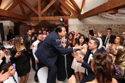 Un homme chante avec un micro au milieu d'une fête, entouré d'invités applaudissant dans une salle décorée.