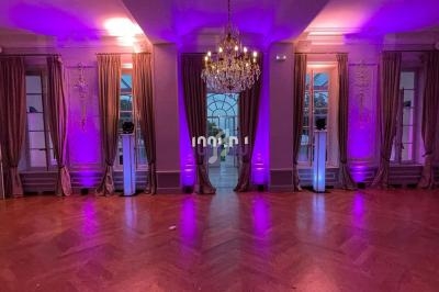 Salle élégante avec parquet en bois, lustre central, rideaux encadrant des fenêtres et éclairage violet diffus.