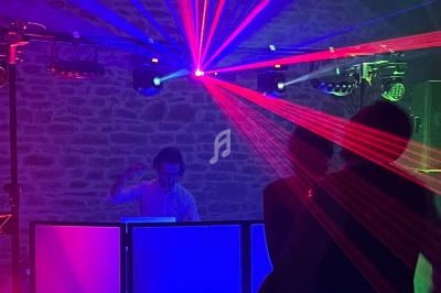 Un DJ mixe devant un mur en pierre, éclairé par des faisceaux lumineux rouges et bleus, avec des silhouettes dansantes au…