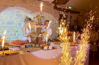 Un couple coupe un gâteau de mariage entouré de desserts et de fontaines lumineuses dans une salle en pierre.