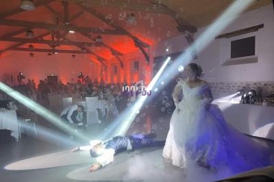 Un couple en tenue de mariage danse sous des lumières et des bulles dans une salle décorée, entouré d'invités.