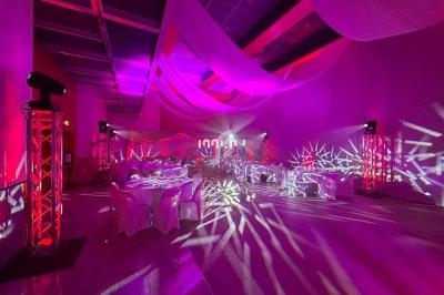 Salle de réception décorée avec des éclairages roses et violets, tables rondes dressées et drapés suspendus.