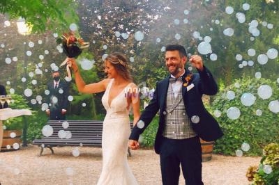 Un couple de mariés souriants marche main dans la main dans un jardin, entouré de bulles et de verdure.